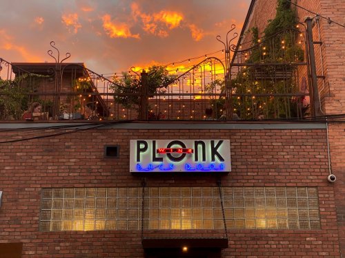 A brick building with a neon sign reading "PLONK" and a rooftop patio, set against a vibrant sunset sky.
