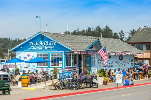 A blue seafood restaurant with outdoor seating, people dining, and an American flag, surrounded by trees and a clear sky.
