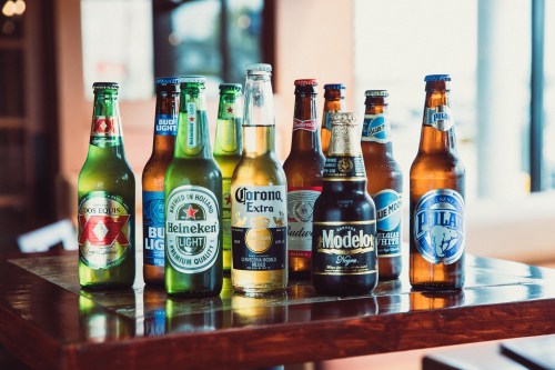 A variety of beer bottles and cans arranged on a wooden table, including brands like Heineken, Corona, and Modelo.