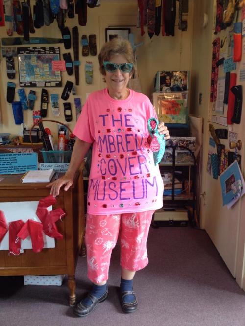 A woman in a bright pink shirt and pants stands in a shop filled with colorful items, holding a teal object.