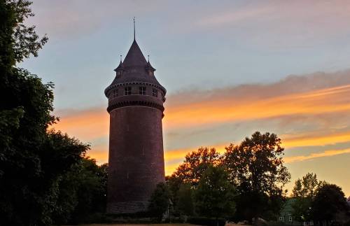 A tall, historic tower silhouetted against a colorful sunset with trees in the foreground.