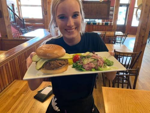 A smiling waitress holds a plate with a burger and a side salad in a cozy restaurant setting.