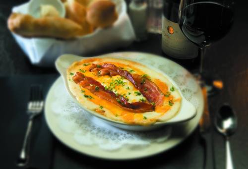A creamy dish topped with bacon and herbs, served with a glass of red wine and bread rolls in the background.