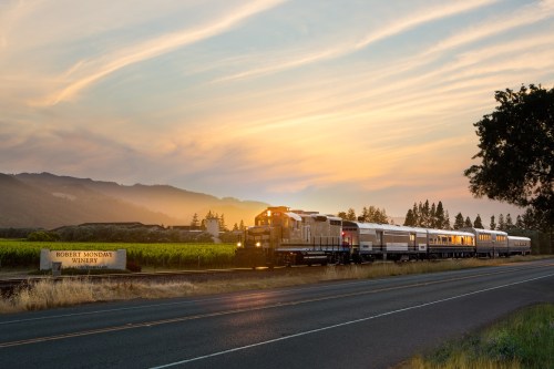 A train passes by a vineyard at sunset, with mountains and a winery sign in the foreground.