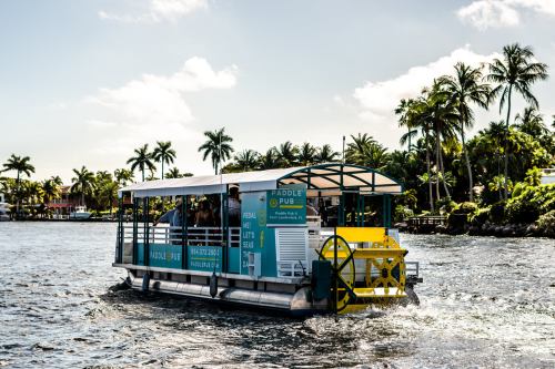A paddle pub boat cruising on a waterway, surrounded by palm trees and sunny skies.