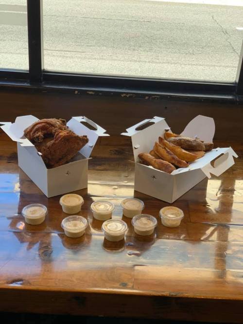 Two takeout boxes with fried chicken and pastries, surrounded by small cups of dipping sauce on a wooden table.