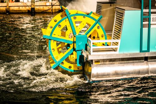 A colorful yellow and turquoise water wheel splashes in the water, part of a larger floating structure.