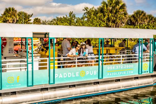 A group enjoying drinks on a colorful paddle pub boat, surrounded by palm trees and water.