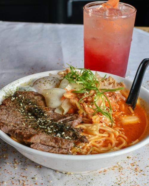 A bowl of spicy ramen topped with beef, green onions, and vegetables, alongside a colorful drink.