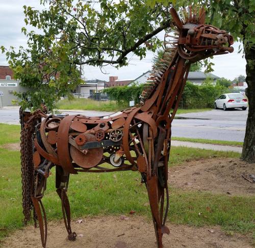 A sculpture of a horse made from rusted metal gears and parts, standing in a grassy area with trees in the background.