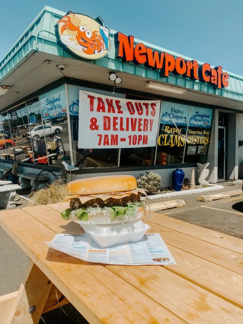 A large sandwich on a table outside Newport Cafe, with a sign for takeout and delivery in the background.