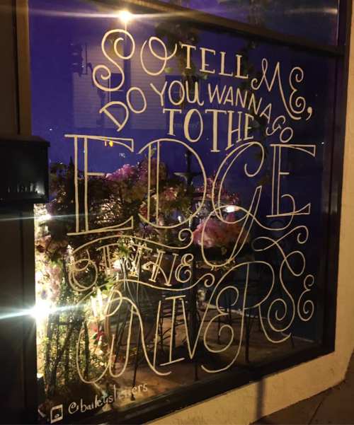 A window display featuring whimsical text: "So tell me, do you wanna go to the edge of the universe?" with floral decor.