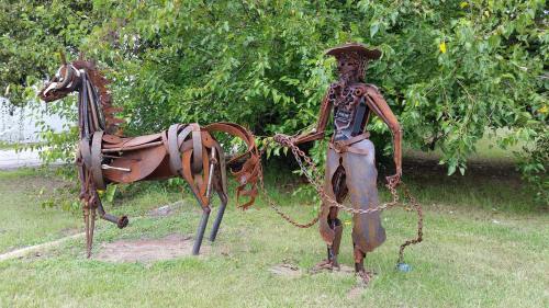 A metal sculpture of a cowboy holding a chain, standing next to a horse made from scrap metal in a grassy area.