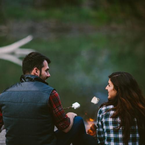 A couple sits by a campfire, roasting marshmallows and enjoying each other's company by a serene lake.
