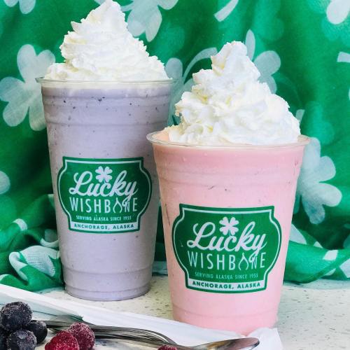 Two colorful milkshakes topped with whipped cream, one purple and one pink, in branded cups against a green background.