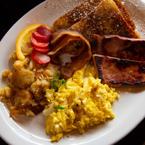 A plate of breakfast featuring French toast, bacon, ham, scrambled eggs, hash browns, and fruit slices.