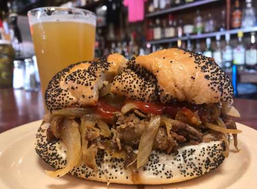 A poppy seed bun filled with beef, sautéed onions, and ketchup, served with a glass of beer in the background.