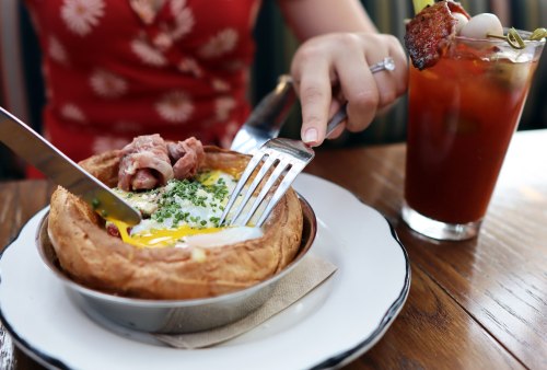 A person cuts into a savory dish with an egg and meat, alongside a cocktail on a wooden table.