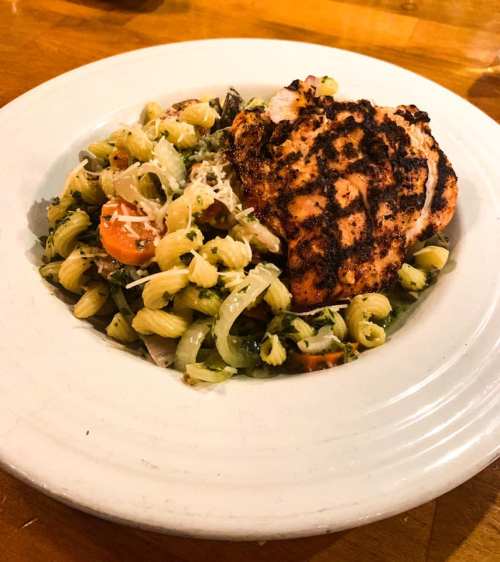 A plate of pasta salad with grilled salmon on top, garnished with cheese and vegetables, served on a wooden table.