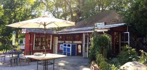 A quaint outdoor bookstore with a patio, surrounded by greenery and featuring tables and umbrellas.
