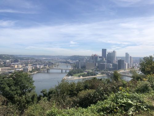 A panoramic view of Pittsburgh's skyline with rivers and greenery in the foreground under a blue sky.