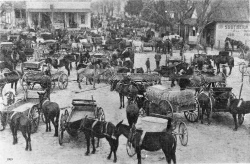 A busy street scene from 1909, filled with horse-drawn carriages and people in a bustling town square.