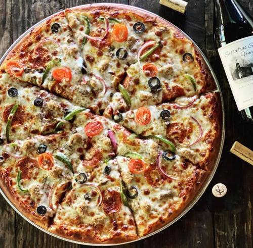 A large pizza topped with pepperoni, olives, bell peppers, onions, and tomatoes, served with wine corks and a bottle.