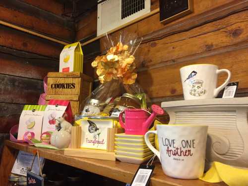 A cozy display of mugs, cookies, and decorative items on wooden shelves, featuring a floral arrangement and colorful packaging.