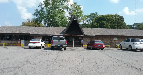 A single-story building with a peaked roof, surrounded by parked cars and trees under a blue sky.