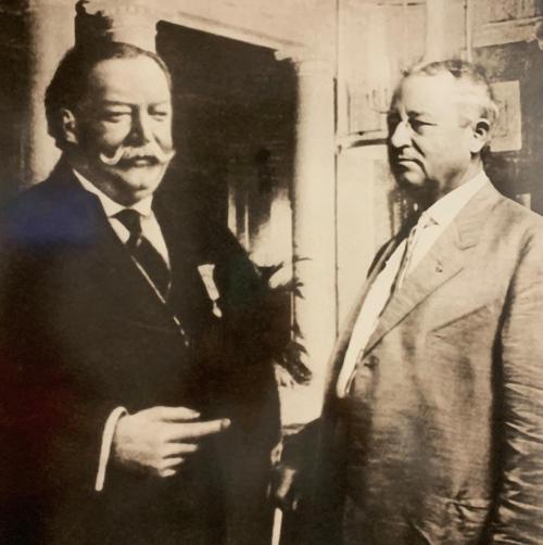 Two men in formal attire, one with a mustache, standing indoors, engaged in conversation. Black and white photo.