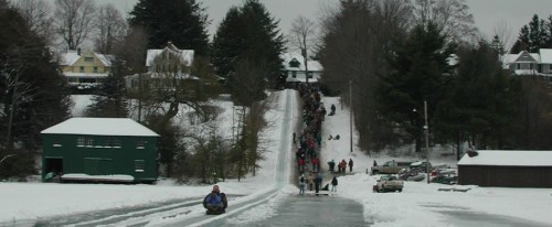 A snowy scene with a long line of people walking up a hill, surrounded by trees and houses, with a frozen lake in the foreground.