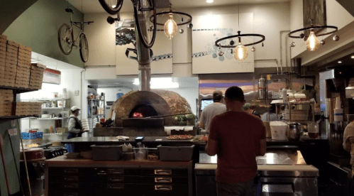 A cozy restaurant kitchen with a wood-fired oven, hanging lights, and a person preparing food at the counter.