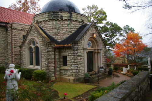 A stone building with a dome, surrounded by greenery and autumn trees, featuring a small entrance and statues.