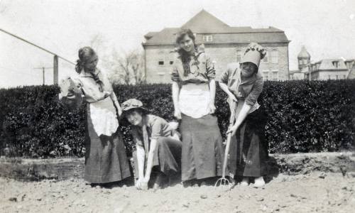 Four women in vintage clothing gardening, with a building and hedge in the background.