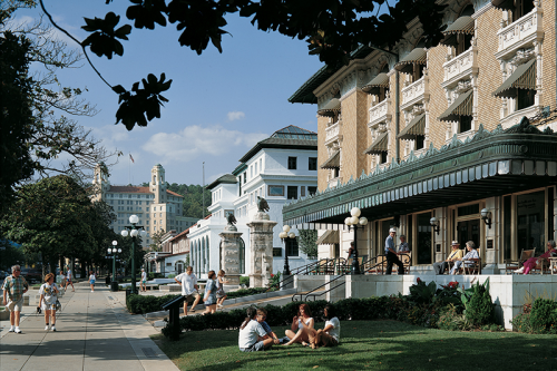 A sunny street scene with people walking and sitting near historic buildings and lush greenery.
