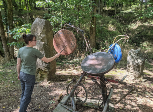 A person strikes a large metal gong in a wooded area, surrounded by sculptures and natural scenery.