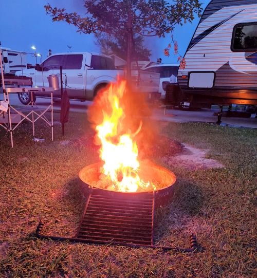 A campfire burns brightly in a fire pit, surrounded by grass and a camping setup with a trailer in the background.