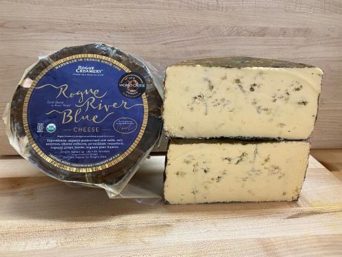 A round wheel of Rogue River Blue cheese next to two cut pieces, showcasing its creamy texture and blue veins.
