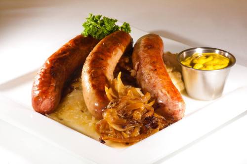 Three grilled sausages served with sautéed onions, sauerkraut, and a side of mustard on a white plate.