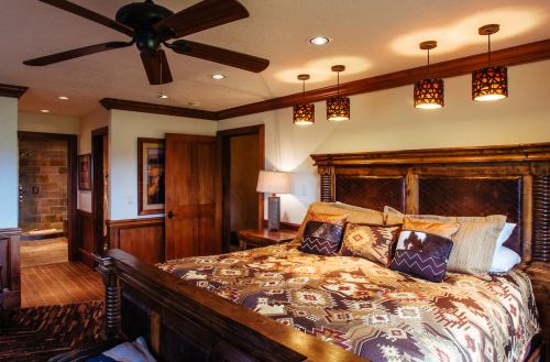 Cozy bedroom featuring a wooden bed, decorative pillows, ceiling fan, and warm lighting. Rustic decor with earthy tones.