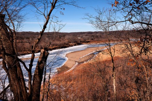 A serene landscape featuring a riverbank with patches of ice and sand, surrounded by bare trees and rolling hills.