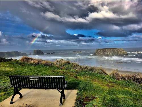 A scenic view of a coastline with a bench, ocean waves, rocky formations, and a rainbow under a cloudy sky.