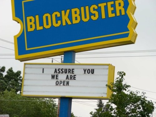 A Blockbuster sign reads, "I assure you we are open," against a cloudy sky and green trees.
