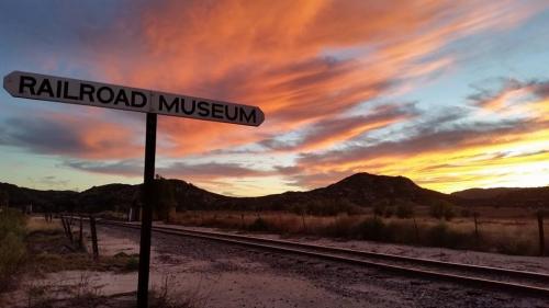 Sign for "Railroad Museum" with a vibrant sunset and mountains in the background, along a railway track.