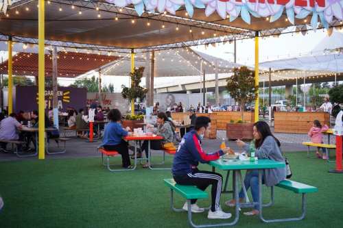 People dining at outdoor tables in a vibrant food garden, surrounded by greenery and festive decorations.
