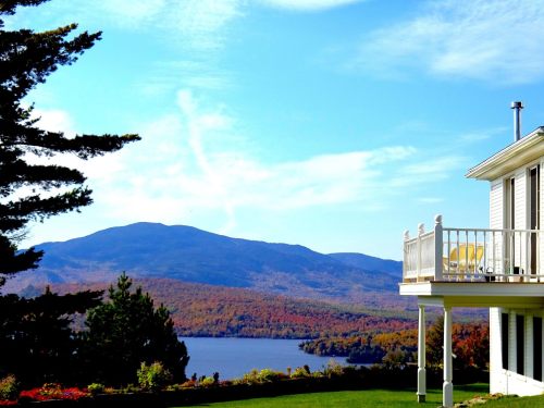 A scenic view of a lake and mountains in autumn, with a white house and balcony in the foreground.