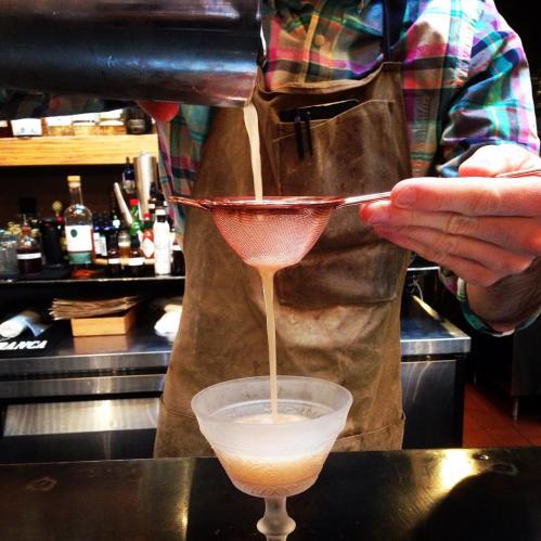 A bartender strains a cocktail into a glass using a fine mesh strainer, with bottles in the background.