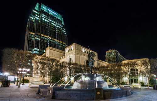 Night view of a fountain with a statue, surrounded by buildings and trees, illuminated by city lights.