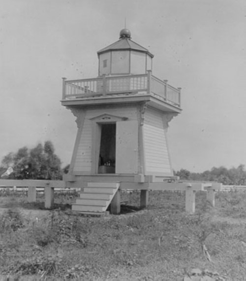 A historic lighthouse with a square base, balcony, and dome, surrounded by grass and trees in a black-and-white photo.