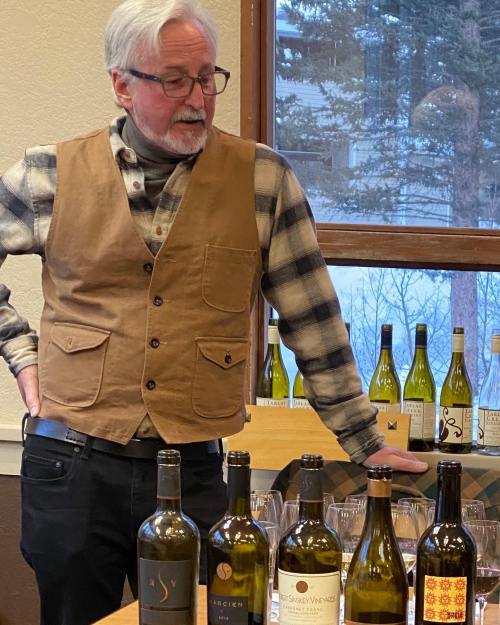 A man in a brown vest and plaid shirt stands by a table with various wine bottles, looking thoughtfully at them.